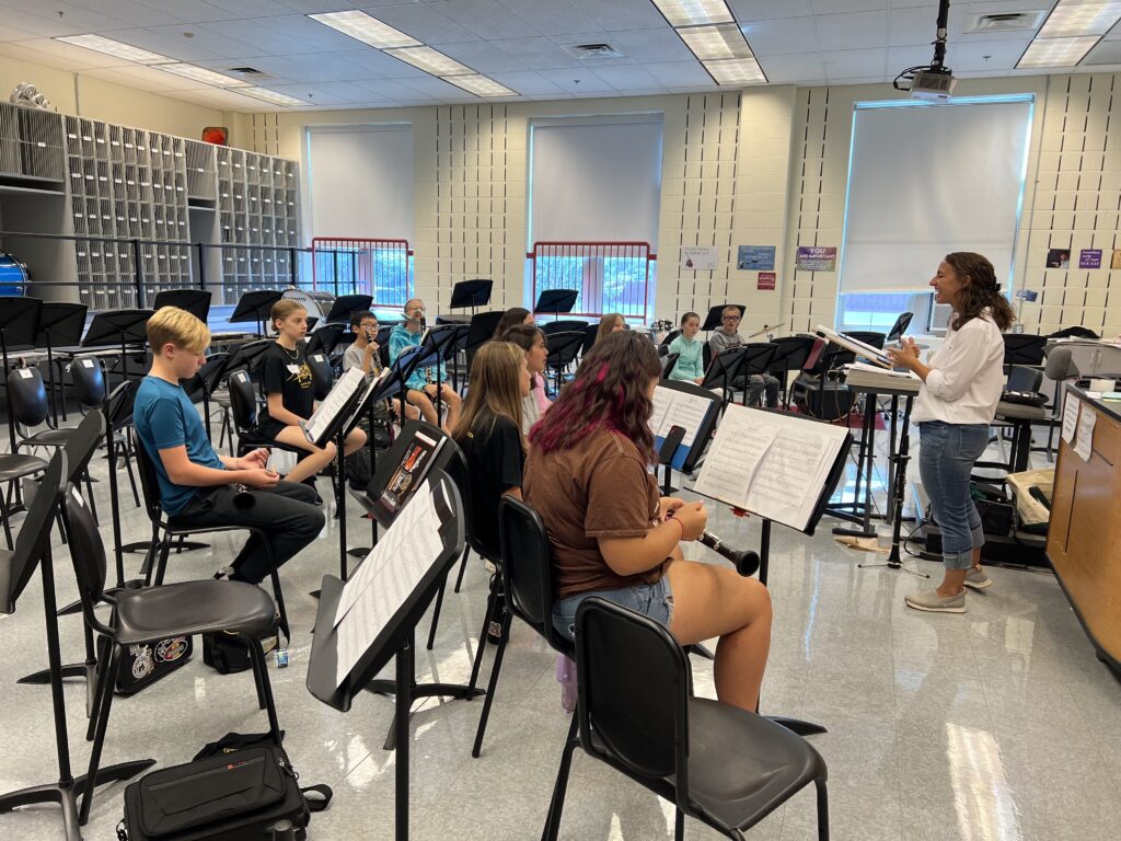 Musicians sit in chairs and look at music on stands while holding clarinets.