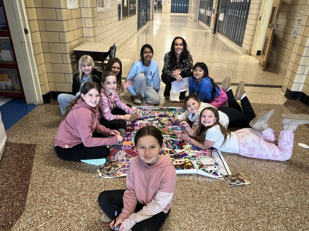 Jamie sits on the floor in the hallway with students as they color a large piece of paper with her designs on it