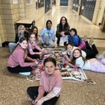 Jamie sits on the floor in the hallway with students as they color a large piece of paper with her designs on it